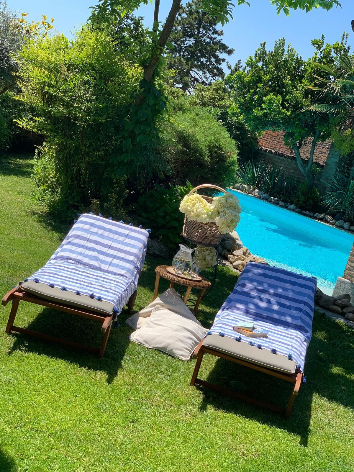 Dos sillones de relax con cojines a rayas, una mesita con una jarrón y vasos, y una cesta de flores junto a una piscina.