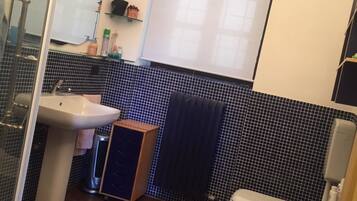 Un baño con lavamanos, inodoro y bidé. Hay un espejo, un perchero para toallas y una ventana con persianas.