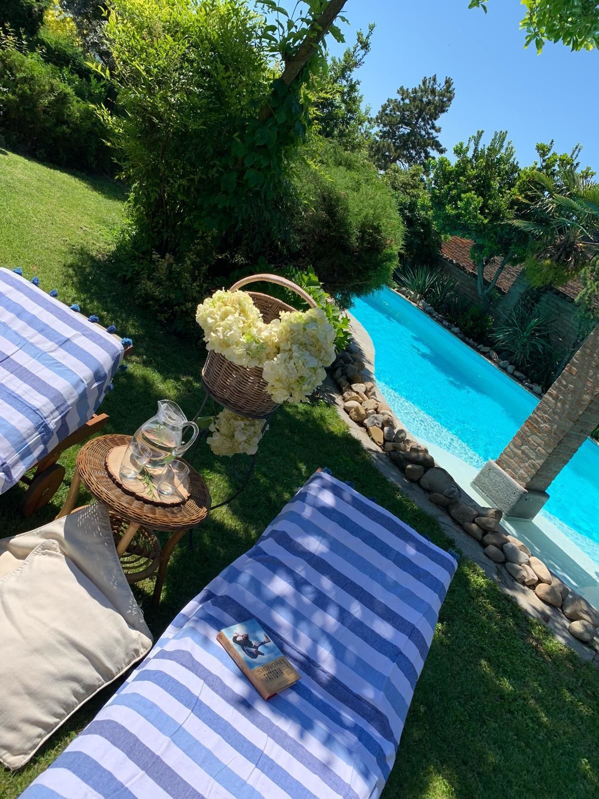 Un espace aménagé au bord de la piscine, avec un fauteuil de relaxation à rayures bleues, une table en osier sur laquelle sont posés une carafe et des verres, et un panier rempli de fleurs blanches.