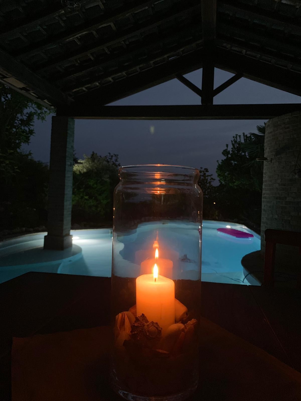 A candle in a jar on a table with a view of a swimming pool at night.