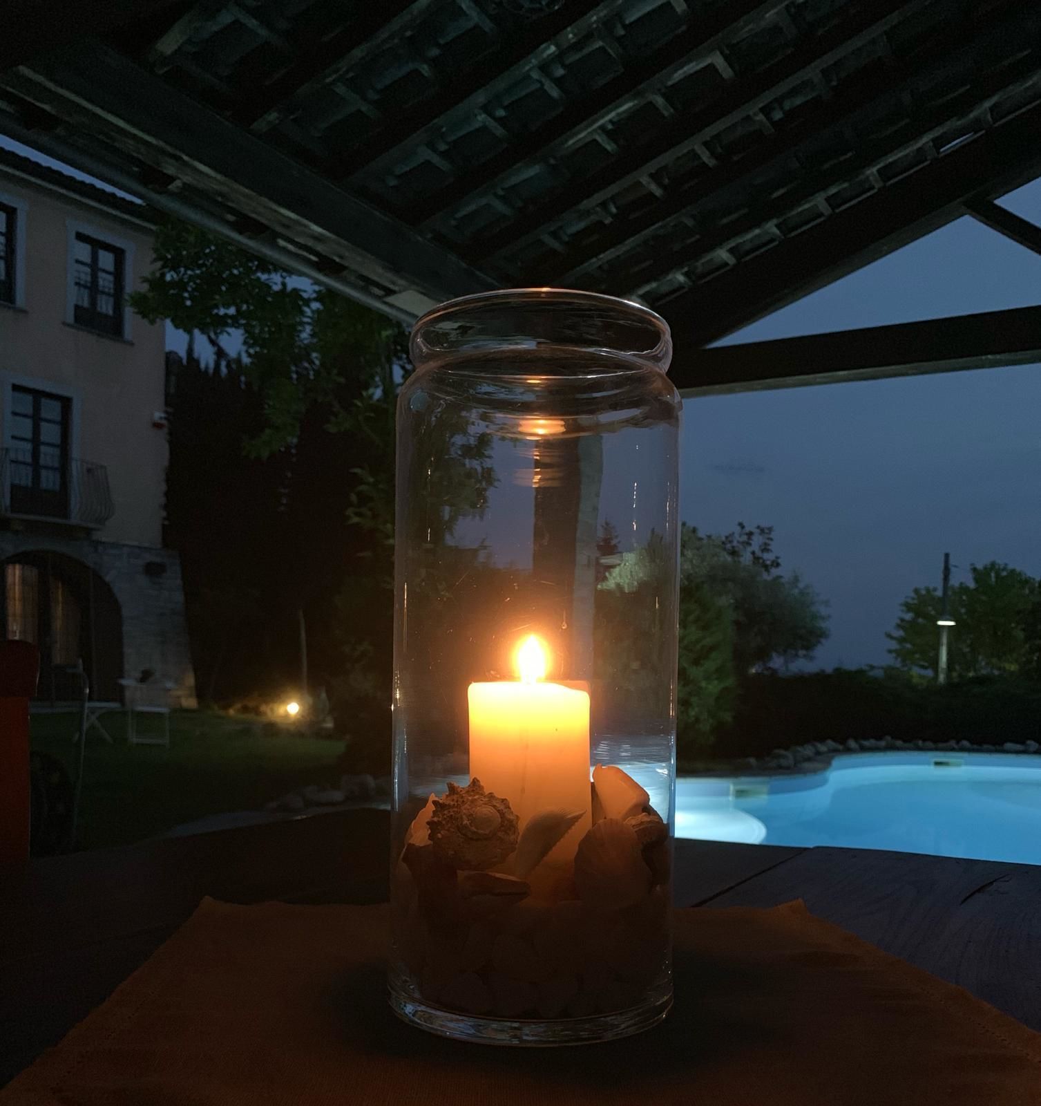 Et stearinlys i et glas med sten, placeret på et bord under et overdækket område med udsigt til en swimmingpool og en bygning i baggrunden.