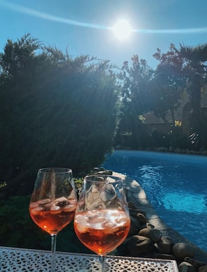 Dos vasos de bebida roja sobre una mesa junto a una piscina, con vistas claras a la piscina y la vegetación circundante.