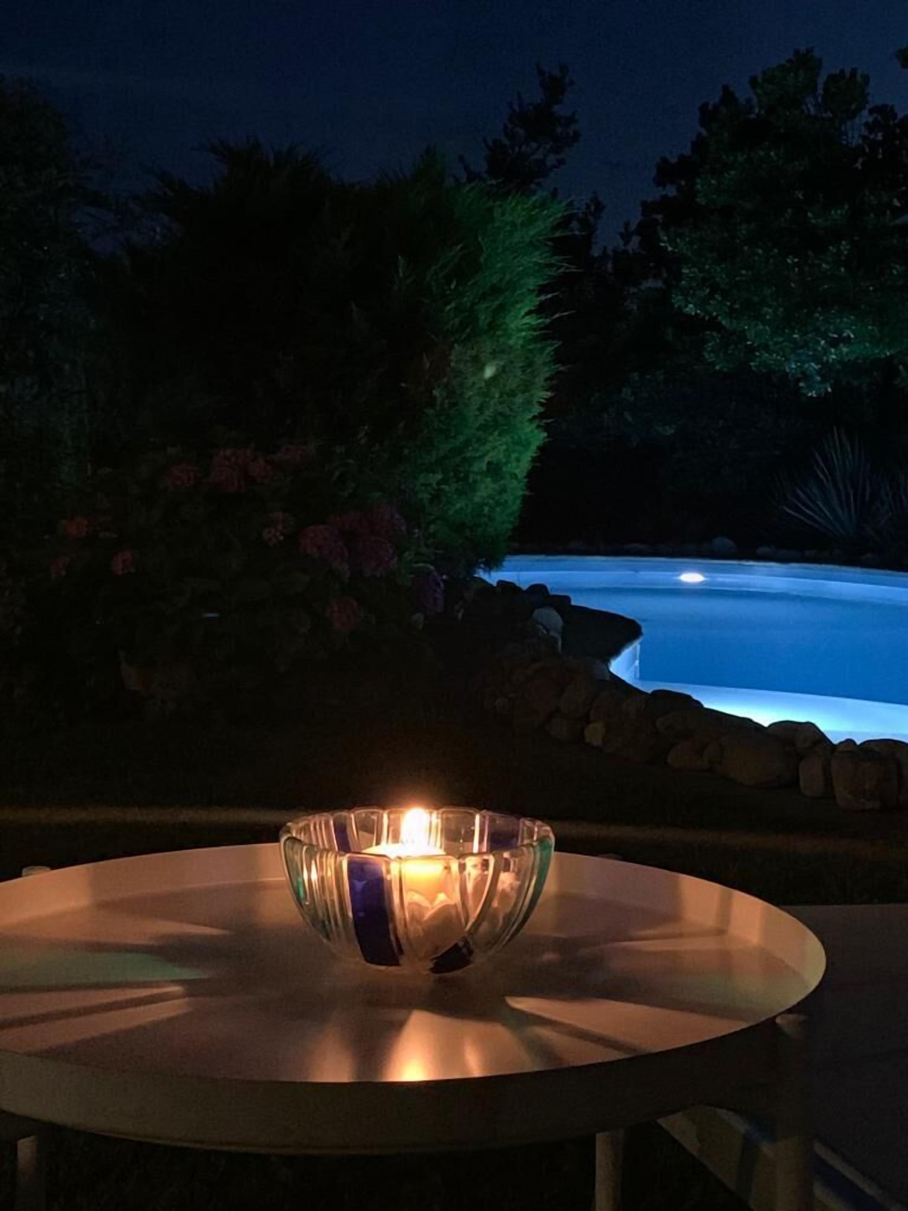 Mesa redonda com uma vela numa taça de vidro, rodeada por um jardim com uma piscina ao fundo.