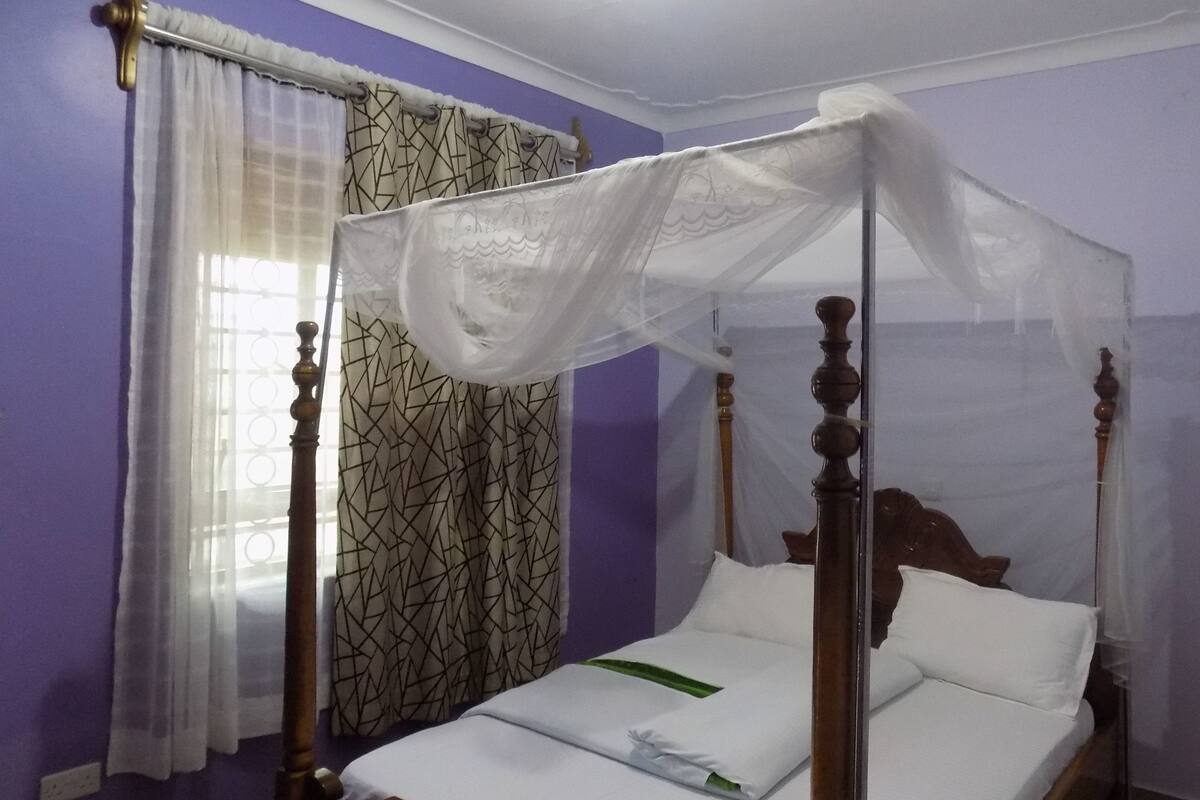 A four-poster bed with a canopy, a window with patterned curtains, and a purple wall.
