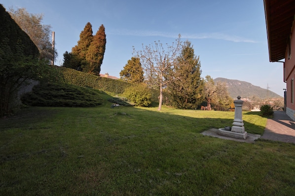 A well-maintained garden with a stone monument, neatly trimmed hedges, and a clear view of a mountain in the background.