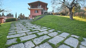 A stone pathway leading to a pink building with a balcony, surrounded by greenery and trees. - Appartamento Il Moresine (Roe Volciano)