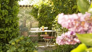 Um pátio com jardim, mesa e cadeiras, cercado por vegetação exuberante e um mural na parede.