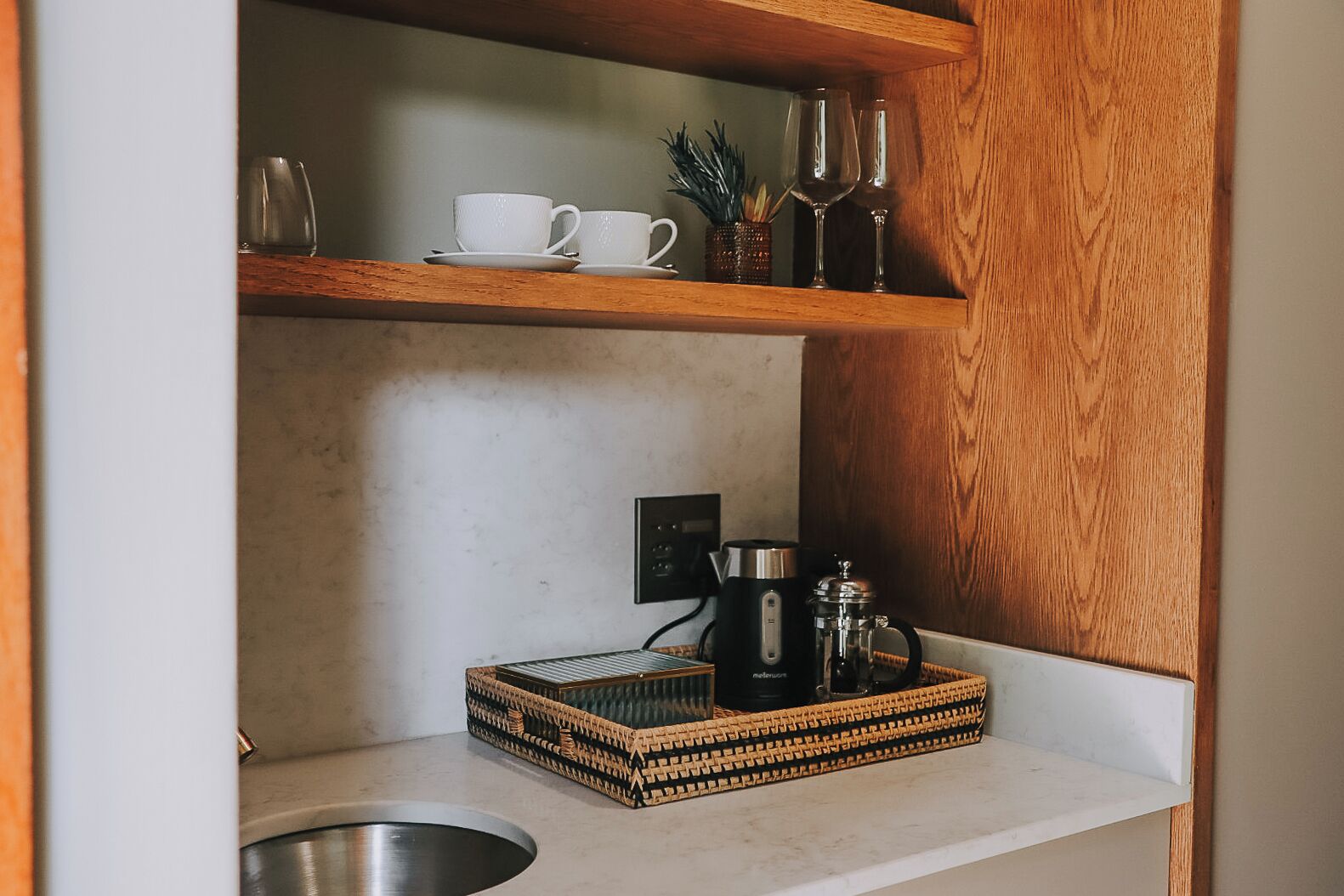 A kitchen with a wooden shelf holding white cups and glasses, a coffee maker, and a decorative tray.