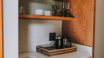 A kitchen with a wooden shelf holding white cups and glasses, a coffee maker, and a decorative tray.