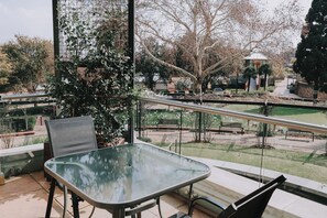 A balcony with a glass table and chairs, overlooking a park with benches and a gazebo.