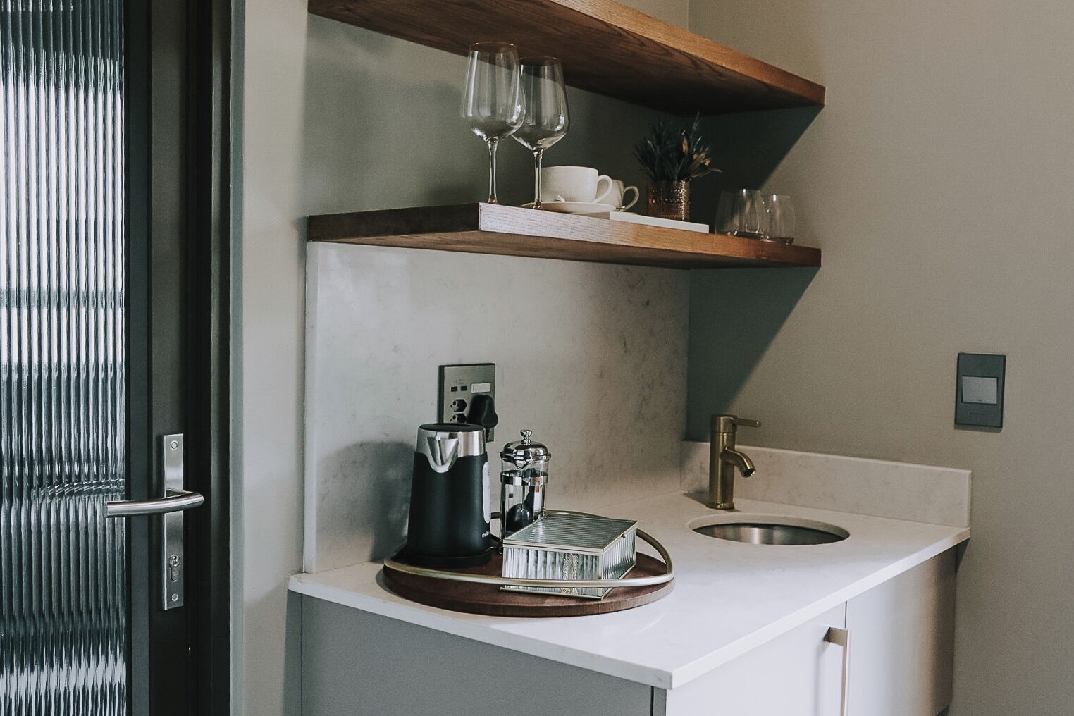 A modern kitchen with a coffee machine, a sink, and a shelf with wine glasses and a cup.
