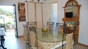 A dining area with a glass table surrounded by wicker chairs, a decorative wall hanging, and a wooden cabinet with a television. - El Garrotal Rural Cottage (Sevilla)