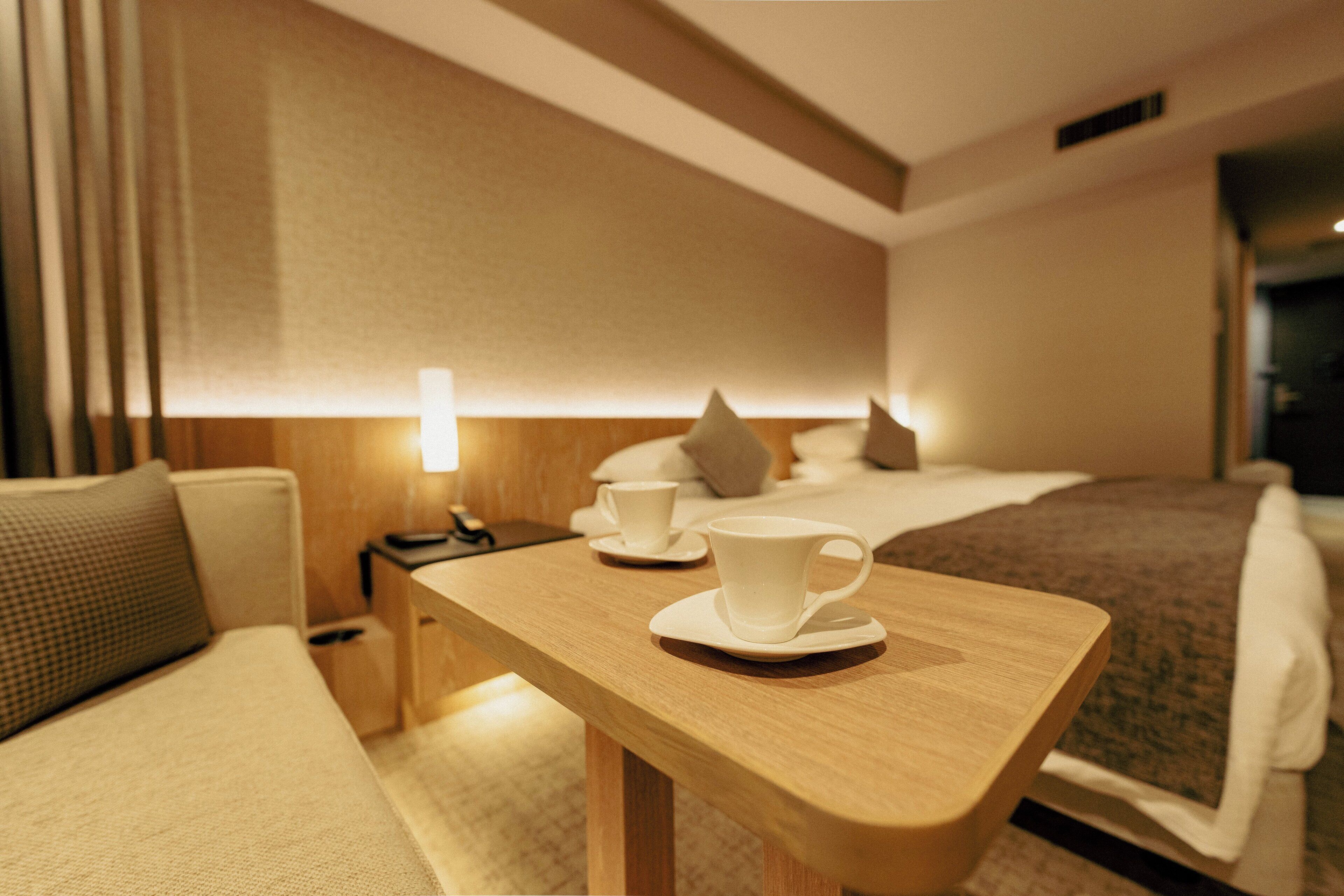 A hotel room with a wooden desk, two white cups, a bed with a brown blanket, and a nightstand with a lamp.