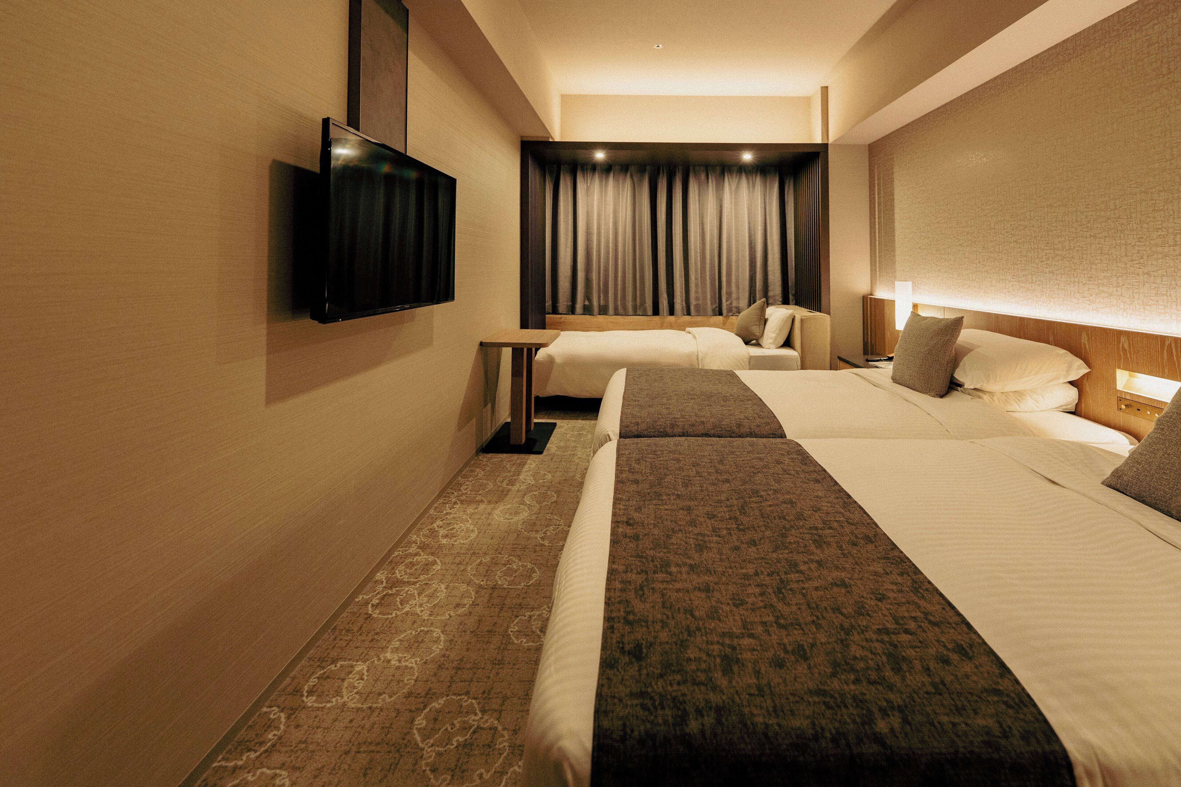 A hotel room with two beds, a flat-screen TV mounted on the wall, a bedside table, and a large window with curtains.