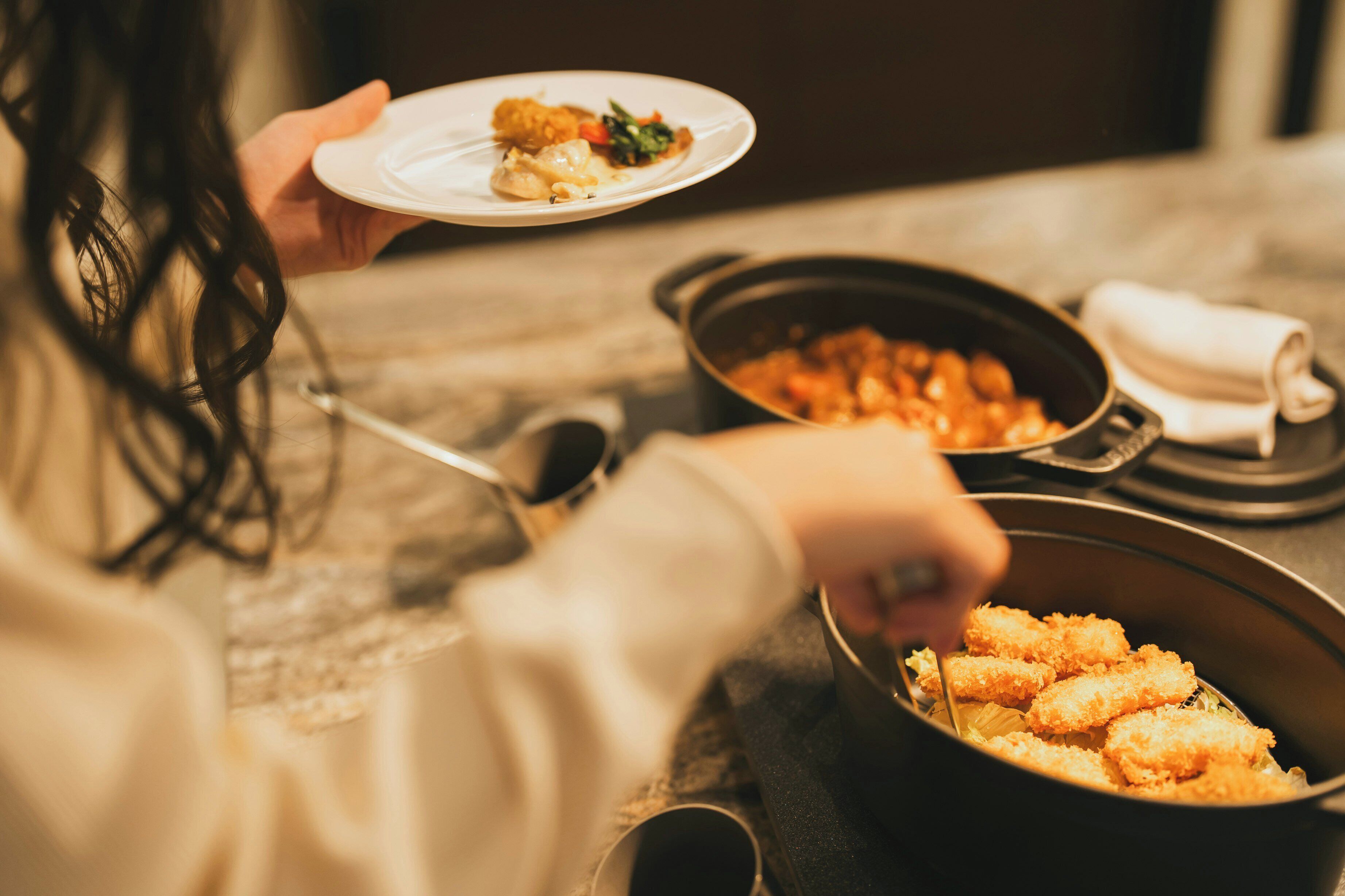 A person holding a plate with food, a pan with a dish, and a bowl with a different dish.