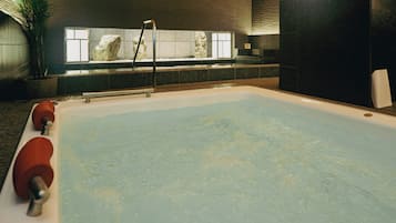 Una amplia piscina interior con superficie de agua cristalina, un tobogán y una pared de piedra con una ventana al fondo.
