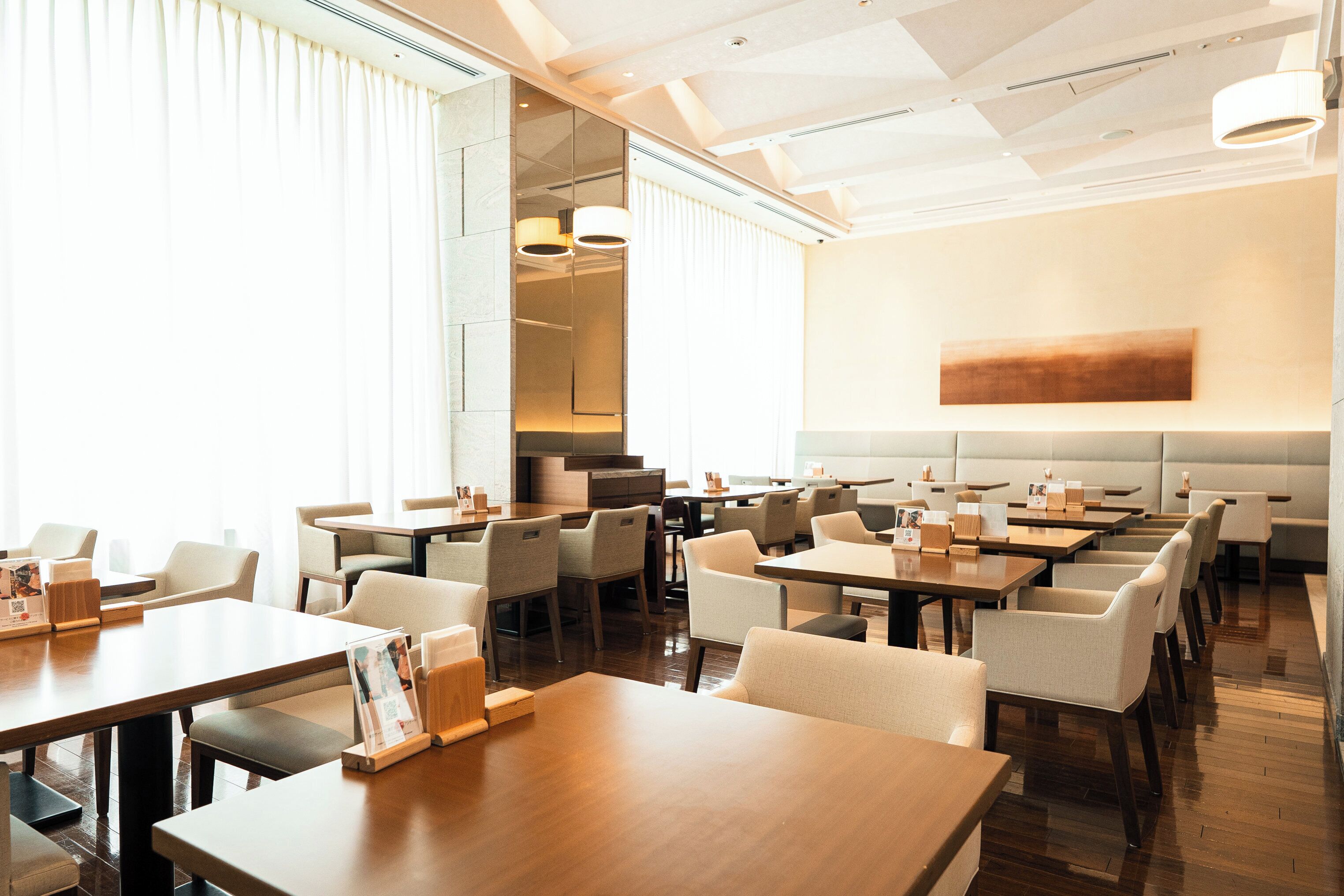 Een ruim restaurant met meerdere tafels en stoelen, grote ramen met zonwering en een modern interieur.