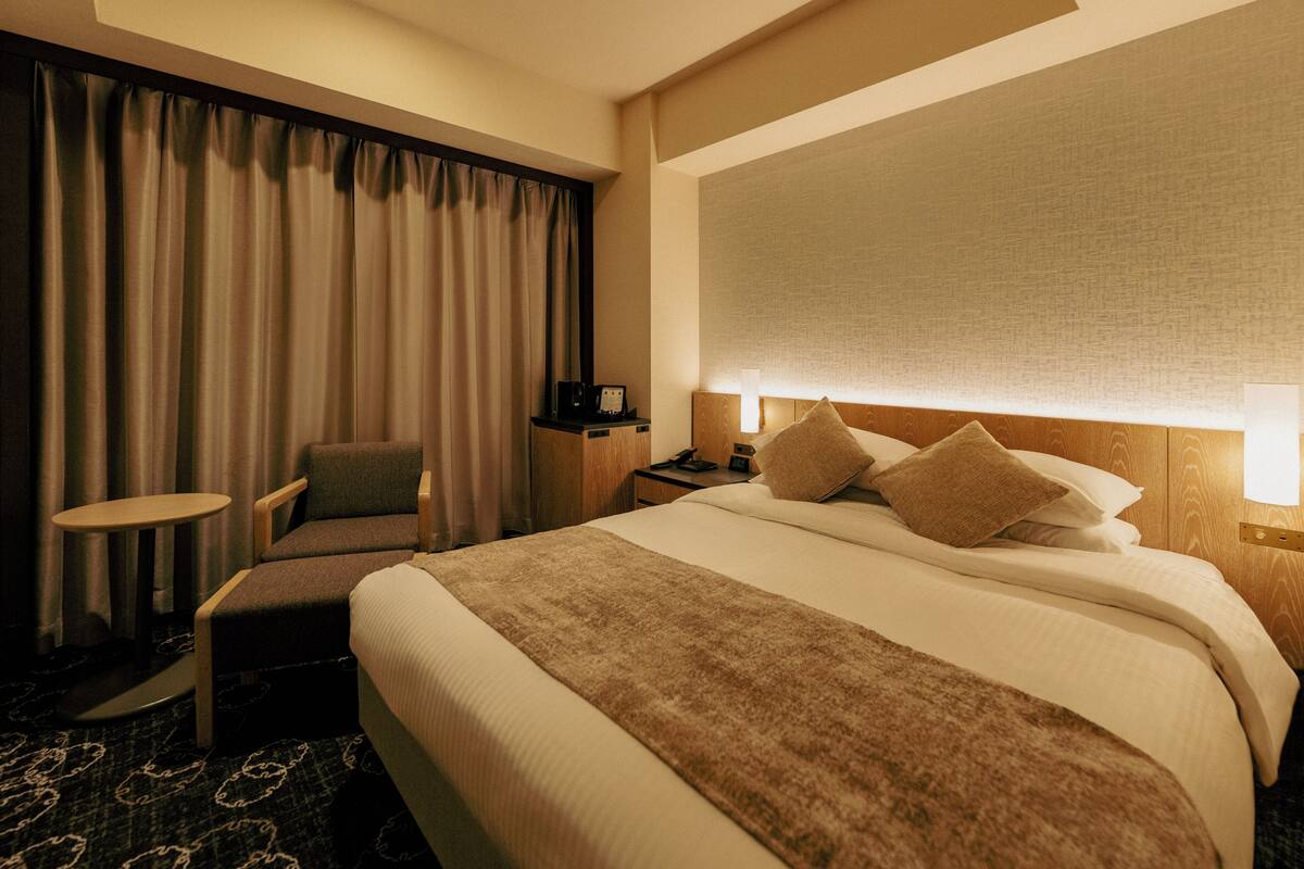 A hotel room with a bed, a chair, a small table, a nightstand, and a window with curtains.
