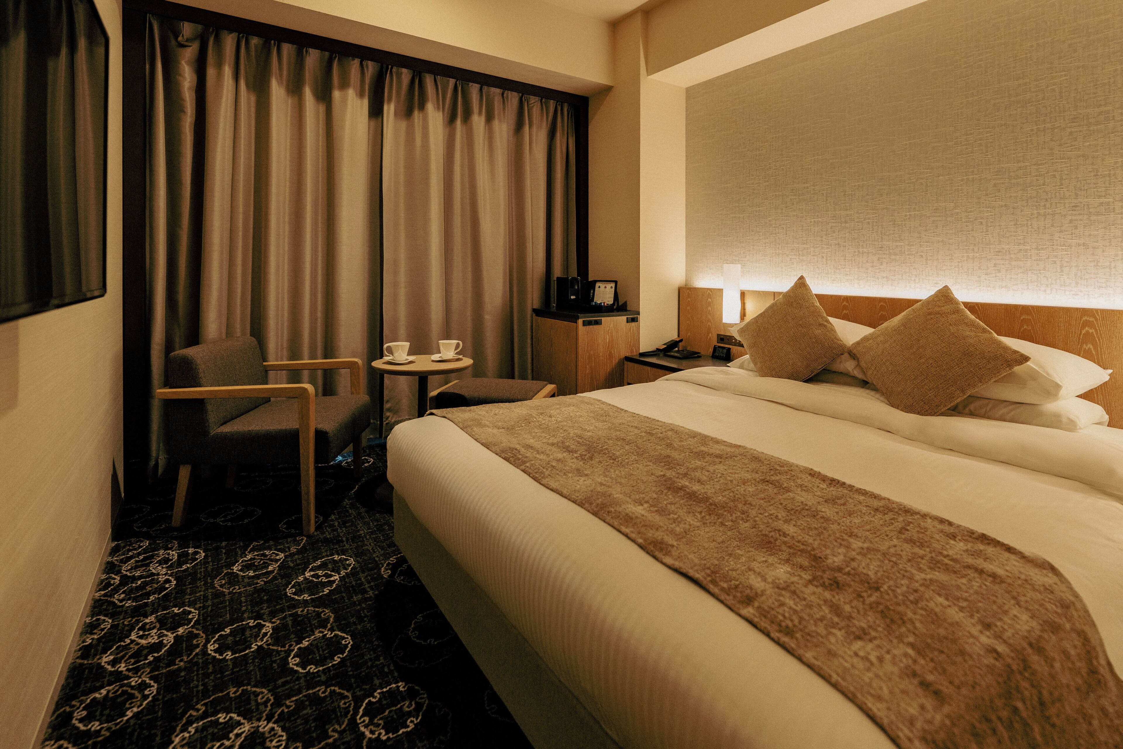 A hotel room with a bed, a desk, a chair, a small table with cups, and a nightstand with a lamp.