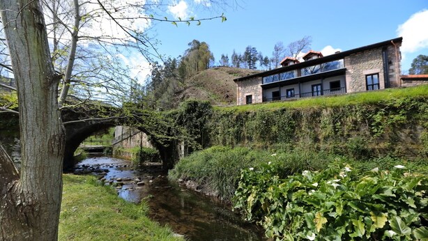 Blick auf einen Fluss mit einer Steinbrücke, üppigem Grün und einem Gebäude mit Steinverkleidung und rotem Dach.