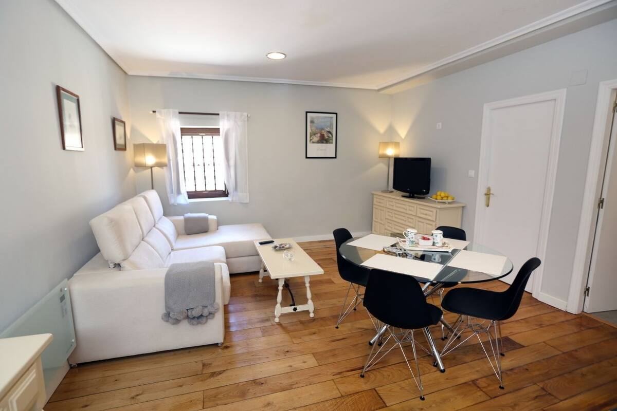 Un espacio habitable compacto con un sofá blanco, una pequeña mesa de comedor con sillas, un televisor y una ventana con cortinas.