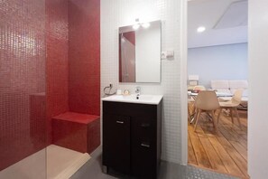 Un baño con ducha de azulejos rojos, un lavabo blanco con espejo y suelo de madera que conduce a un dormitorio con cama y sillas.
