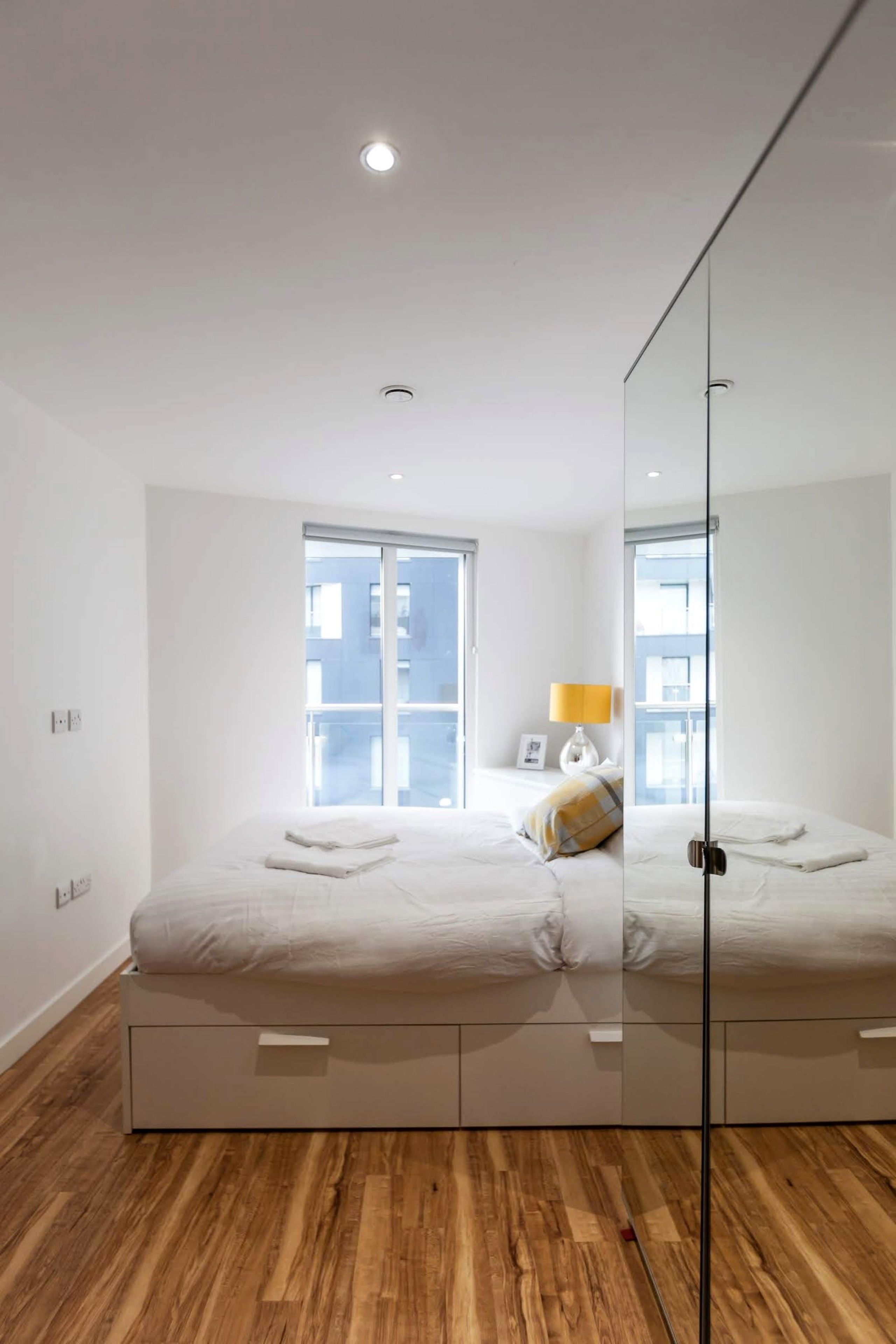 A modern bedroom with a large mirror, a bed with storage drawers, a window with a view of buildings, and a bedside lamp.
