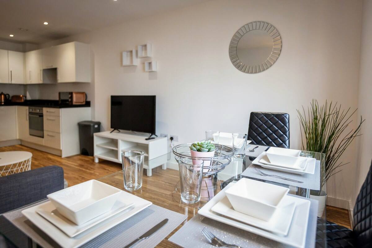 A modern dining area with a glass table set for four, a television, and a kitchen in the background.