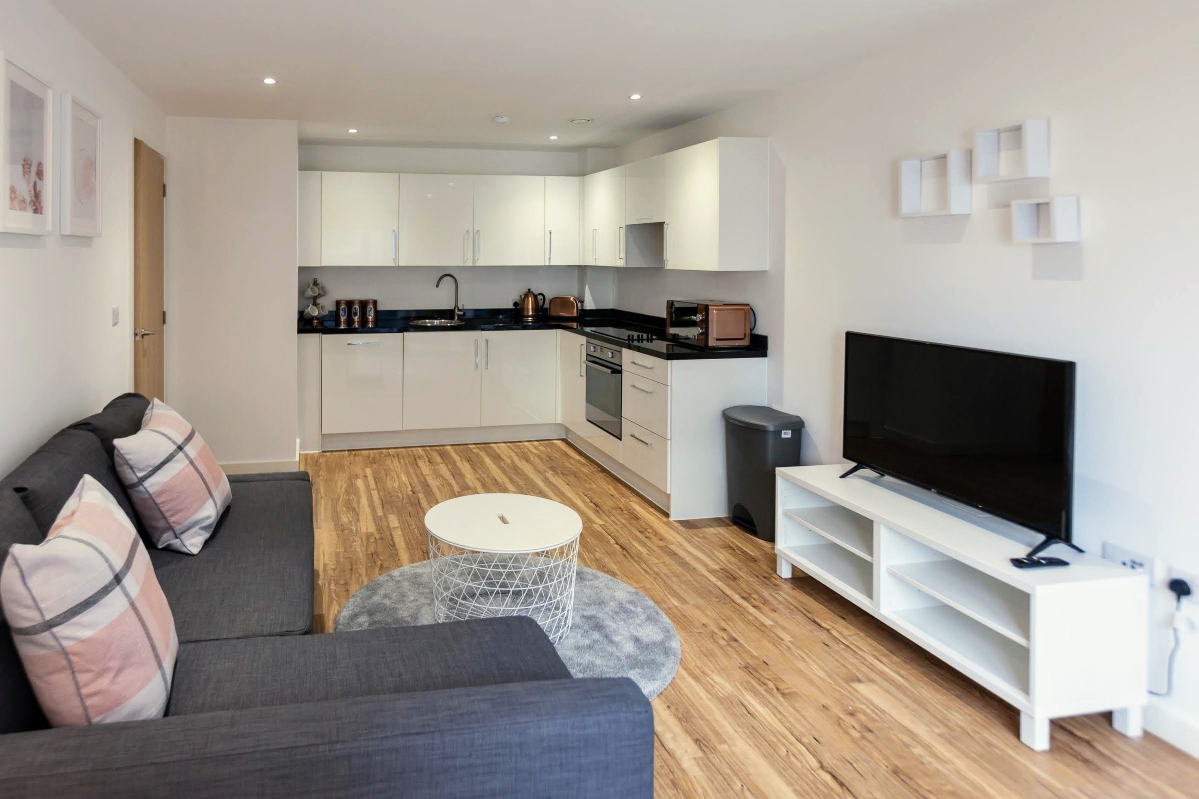 A modern living room with a grey sofa, a coffee table, a TV on a white stand, and a kitchen in the background.