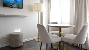 A modern hotel room with a round dining table, two chairs, a tufted ottoman, a floor lamp, and a flat-screen TV mounted on the wall. - Famous Crows Lisbon Suites (Lisbon)