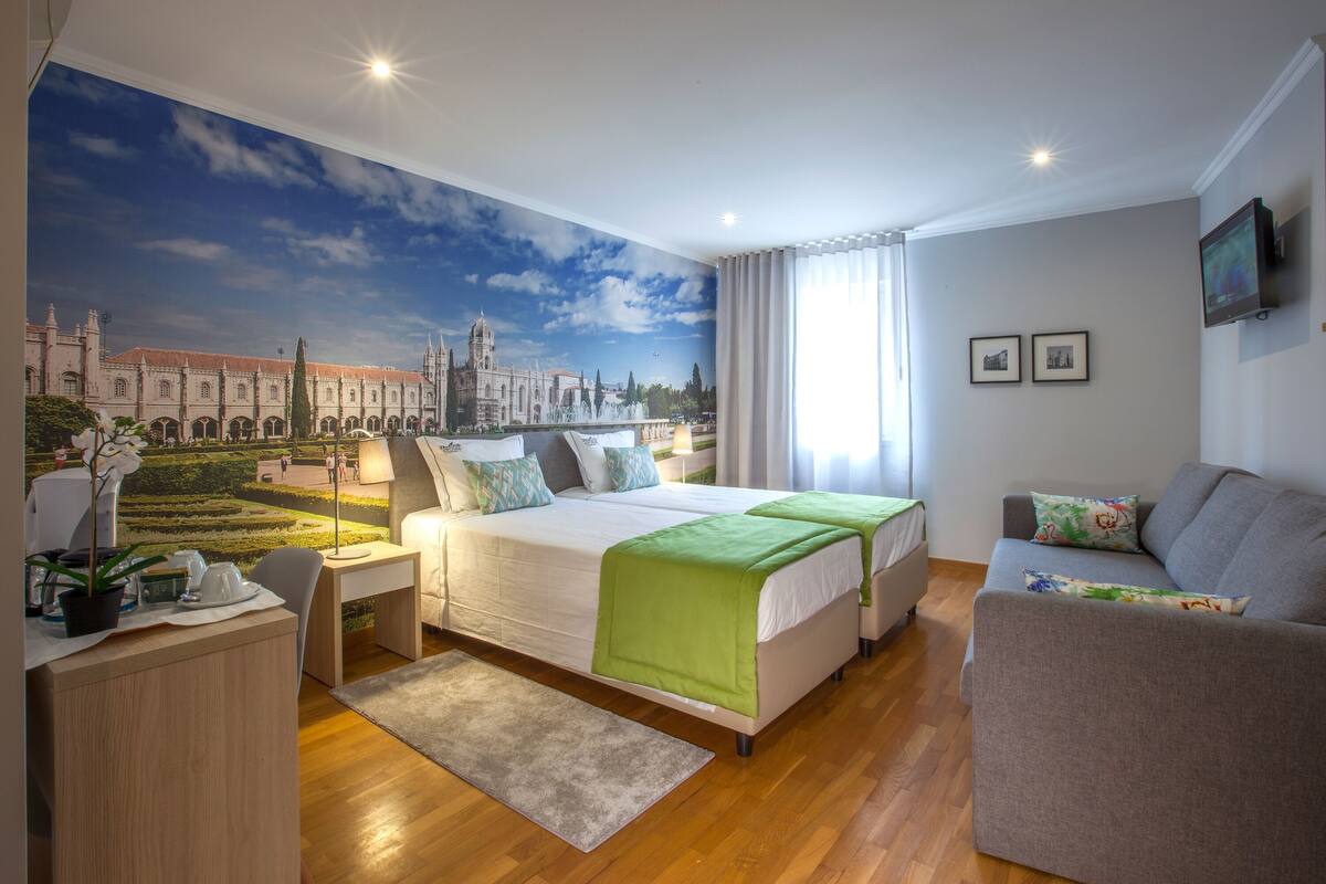 Quarto de hotel com um grande mural de um horizonte urbano, uma cama com roupa de cama verde, um sofá cinzento e uma escrivaninha com uma luminária.