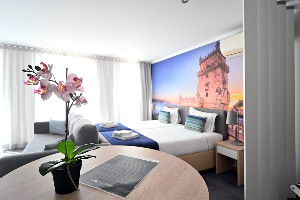 Quarto de hotel com uma cama, uma escrivaninha, uma cadeira, uma planta em vaso e um mural na parede com um castelo.