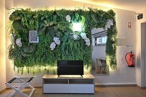 Una habitación con un televisor de pantalla plana sobre un soporte blanco, una silla blanca y una pared decorada con follaje verde y flores blancas.