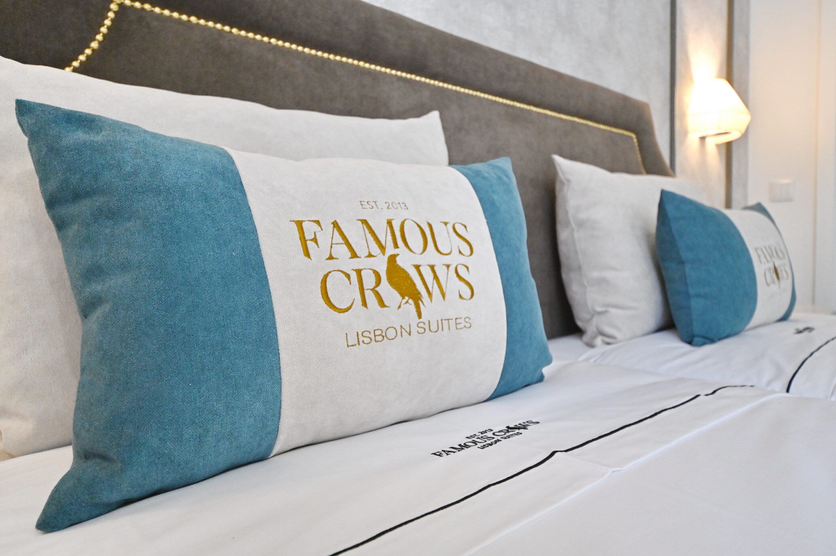 A hotel bed with pillows featuring the logo "Famous Crows Lisbon Suites."