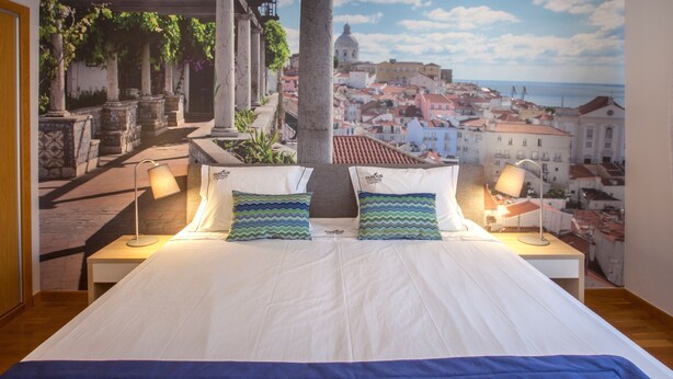 A bedroom with a large bed, two bedside lamps, and a cityscape mural on the wall. - Famous Crows Lisbon Suites (Lisbon)