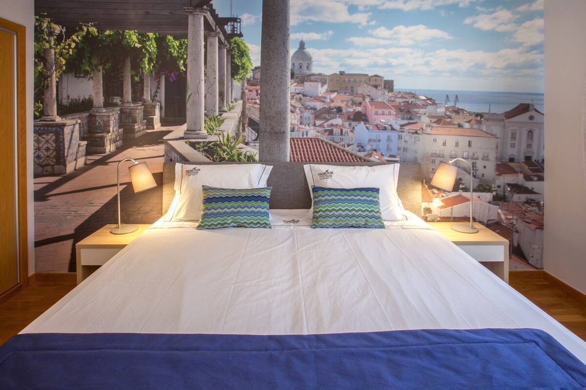 Quarto com uma cama grande, duas luminárias de cabeceira e um mural com vista para a cidade.