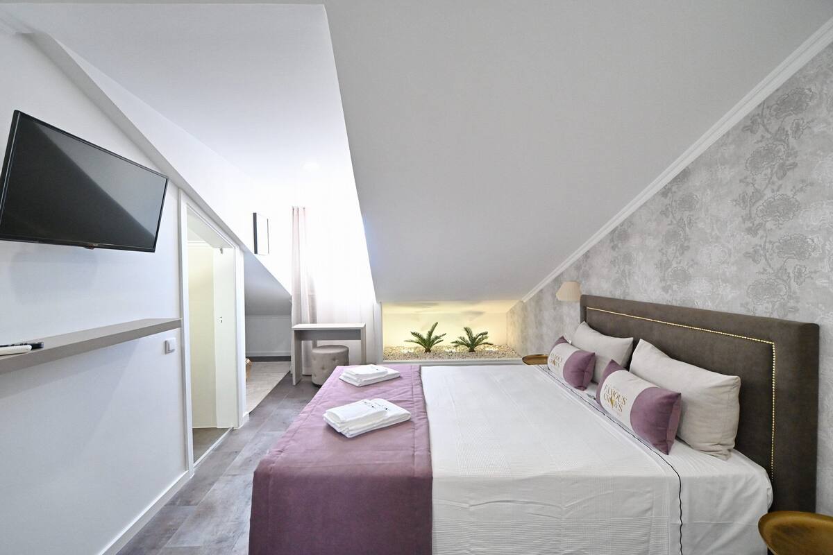 Quarto de hotel moderno com uma cama grande, uma TV de tela plana na parede e vista do céu através de um claraboia.