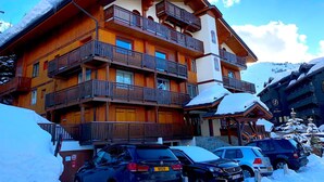 Un edificio de madera de varios pisos con balcones, una pista de esquí al fondo y varios coches estacionados en la parte delantera.