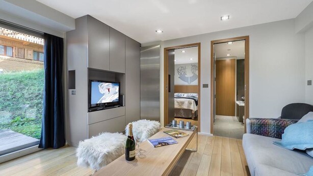 Una sala de estar moderna con un televisor de pantalla plana, una mesa de centro de madera con una botella y vasos, un sofá y vistas a un jardín a través de amplios ventanales.