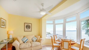 A room with a beige sofa, wooden chairs, a dining table, and large windows offering a view of the sea. - Lovely south facing apartment with breathtaking views of the whole of Torbay. (Torquay)