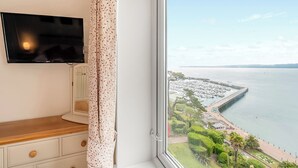 A room with a TV mounted on the wall, a wooden dresser, and a window offering a view of a marina and coastline. - Lovely south facing apartment with breathtaking views of the whole of Torbay. (Torquay)