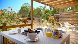 A wooden deck with a table set for breakfast, including croissants, a fruit bowl, and a jug of orange juice. - Camping Bella Vista (Calvi)