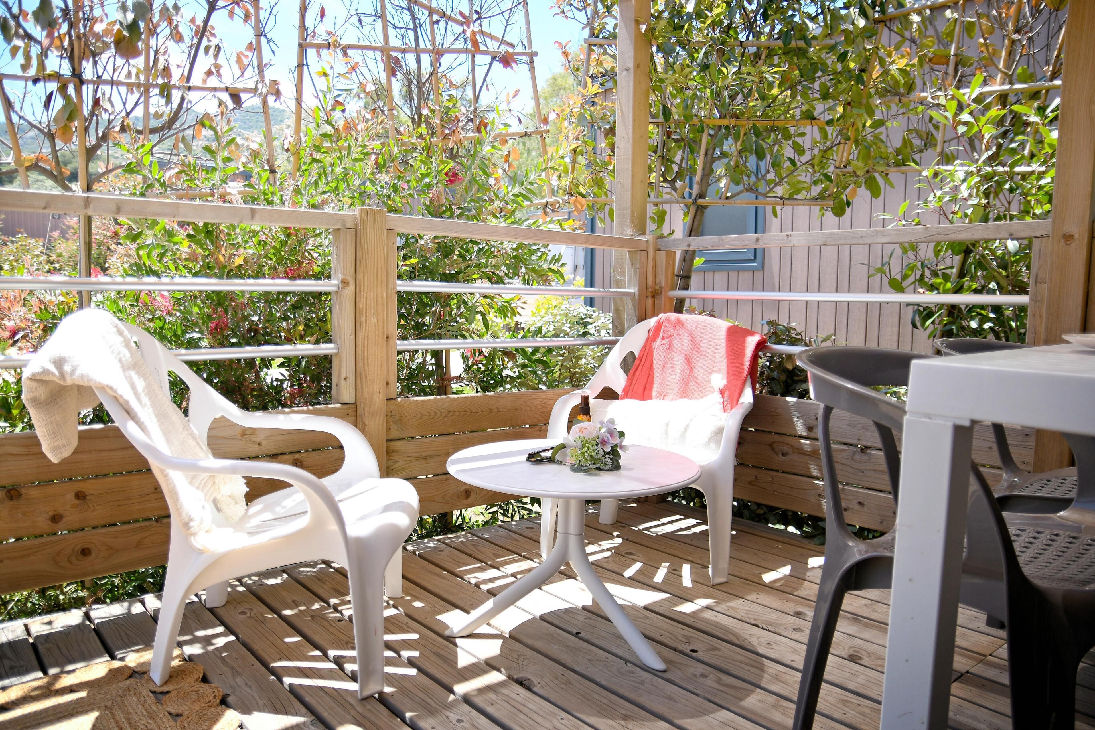 A wooden deck with white plastic chairs and a small round table, surrounded by a wooden fence and greenery.