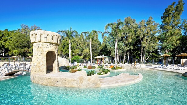 A clear pool with a stone tower, surrounded by lounge chairs and palm trees. - Camping Bella Vista (Calvi)
