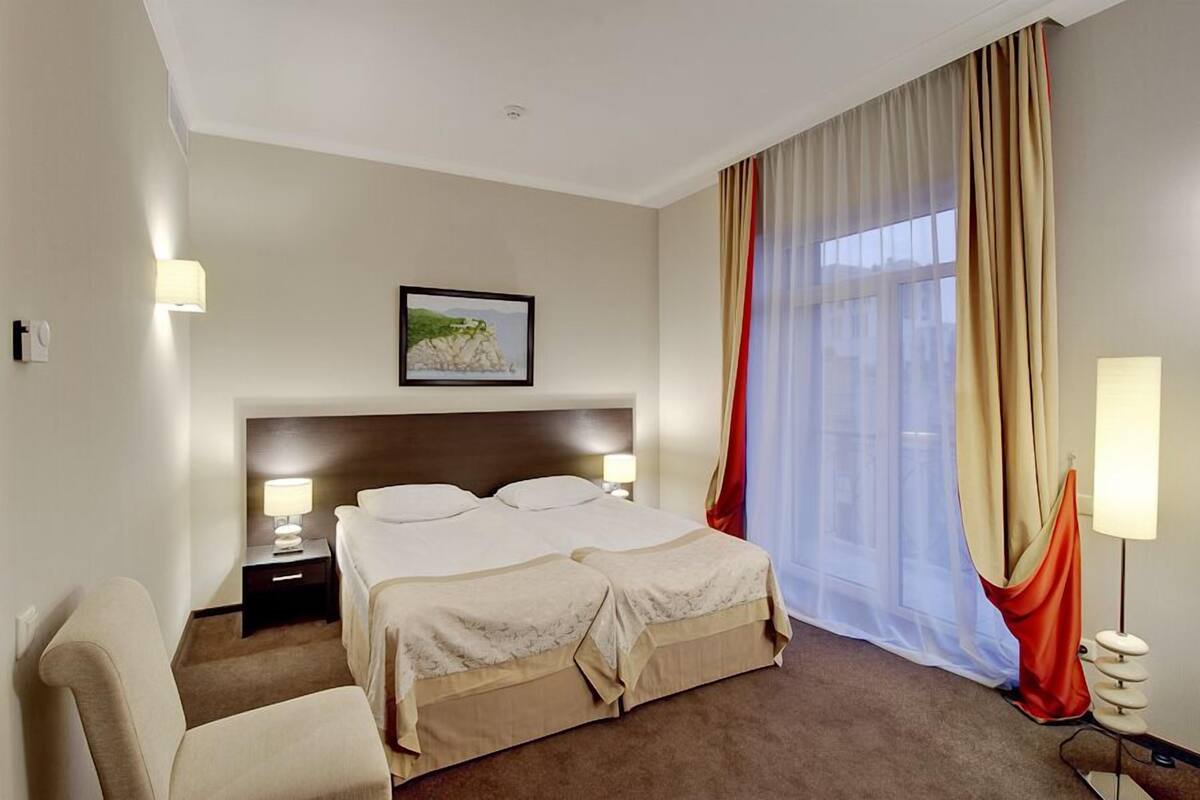 Una habitación de hotel con una cama grande, dos mesitas de noche con lámparas, un cabecero, un cuadro enmarcado en la pared y una ventana con cortinas translúcidas.