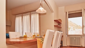 Un coin repas avec une table en bois, des chaises blanches et une nappe jaune. Il y a deux fenêtres avec des rideaux blancs, un radiateur et une étagère remplie de livres.