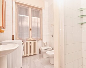 A bathroom with a toilet, bidet, radiator, and a window with sheer curtains. - Appartamento San Giuseppe (Salò)