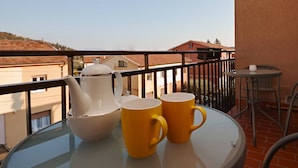 Un balcon avec une table dressée pour deux personnes, une théière blanche et deux tasses jaunes.