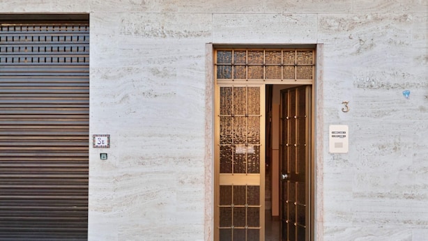 Une entrée d’immeuble avec une porte à volets fermés, une porte numérotée et une petite pancarte sur le mur.