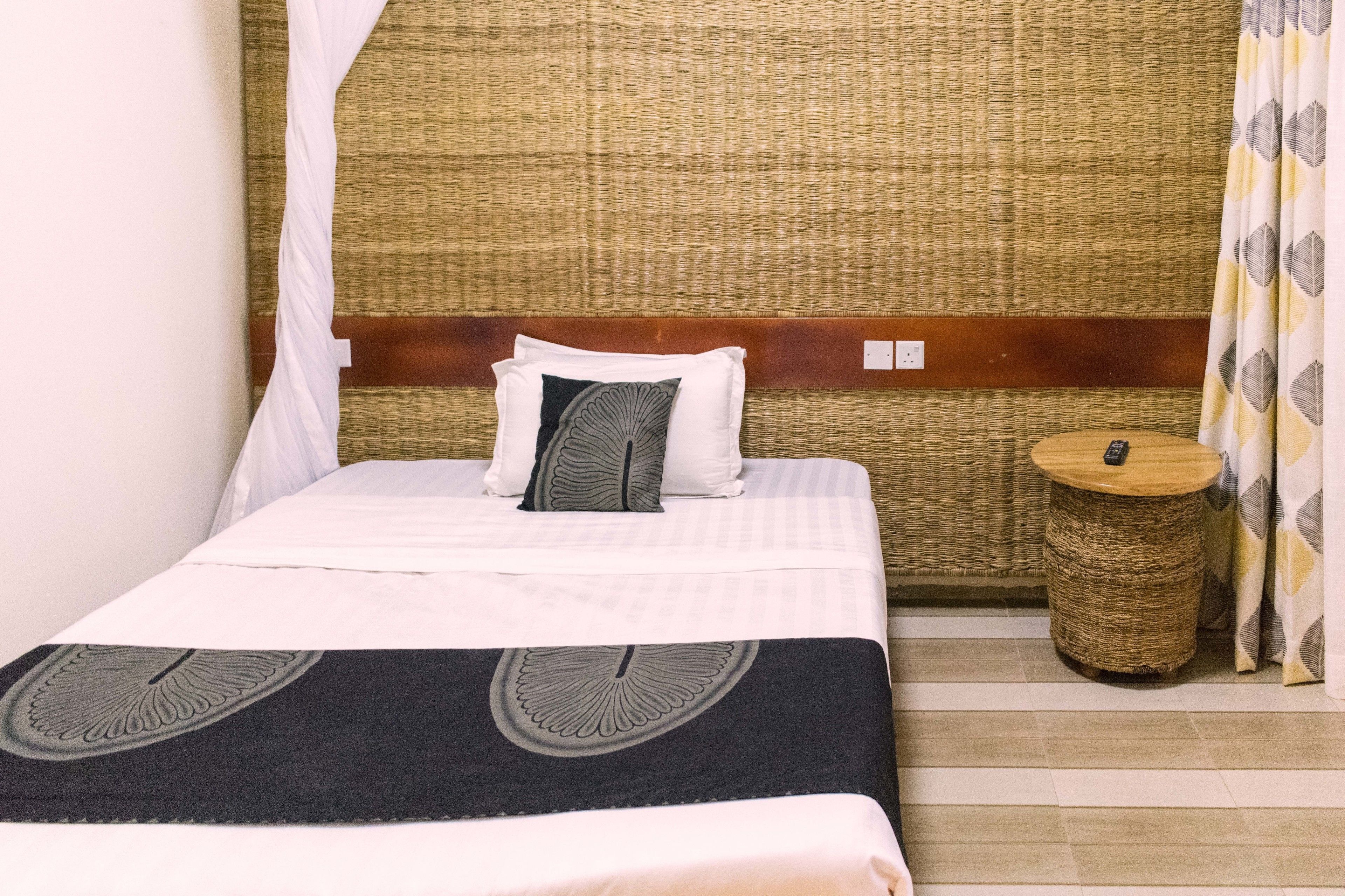 A neatly made bed with a white and black bedspread, a woven headboard, and a small round side table with a remote control.